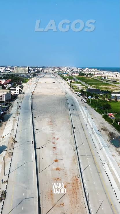 Aerial view of the Coastal Highway under construction through a dense built-up part of Lagos, concrete carriageway laid and the ocean visible in the distance.